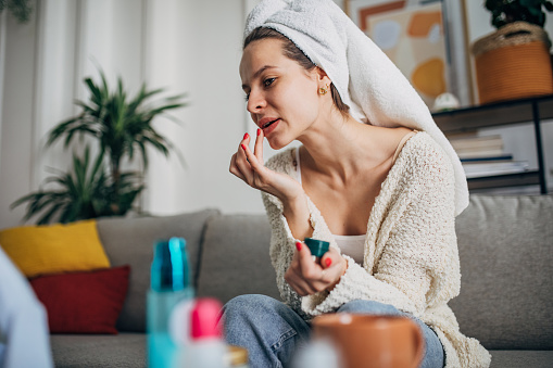 One woman, beautiful young woman sitting on sofa in living room and applying lip balm, she has a towel on her head.