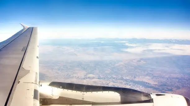 View from the window of an Airbus A321 plane flying above the clouds where we see the engine, a wing and some fields below