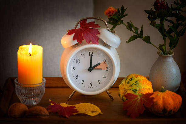 Fall back, time change on a white alarm clock after daylight saving time, candle, leaves and pumpkins as autumn decoration stock photo