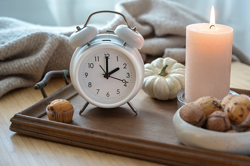 White vintage alarm clock, candle, autumn decoration and sweets on a nightstand with a natural woolen blanket, fall back concept after daylight saving time