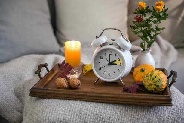 Vintage alarm clock showing one hour fall back after daylight saving time, wooden tray with candle and autumn decoration on a bed with natural blanket and pillows, copy space stock photo