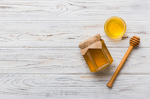 Glass jar of honey with wooden drizzler on colored background. Honey pot and dipper high above. Top view copy space.