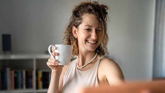 One woman young adult caucasian female standing in the office drinking coffee and using mobile phone to make video call copy space