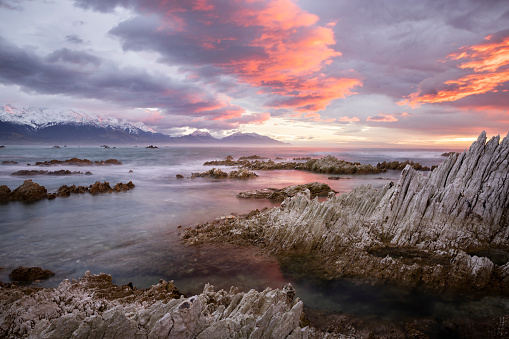 Kaikoura, New Zealand, where the mountains meet the sea. A colorful sunrise lights up the sky over the rocks, ocean and distant snow-capped mountains. Due to a powerful 7.8 magnitude earthquake in 2016, a 20km stretch of the Kaikoura coastline was lifted by up to 2 meters, so the rocks in the foreground would probably have been under water before the quake.