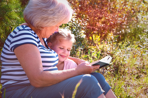 Grandmother reading to her granddaughter child book outdoors. Happy family concept