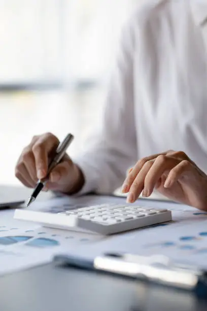 Photo of Businessman is using a calculator to calculate company financial figures from earnings papers, a businessman sitting in his office where the company financial chart is placed.