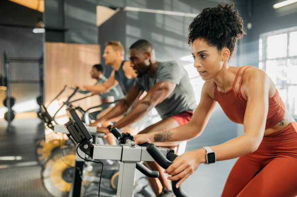 une femme noire avec un groupe sur un vélo d’exercice, faisant du vélo au gymnase pour perdre du poids et améliorer la santé cardiovasculaire. un vélo est la meilleure activité pour le cardio, la perte de graisse saine et l’entraînement pour le  - cours de gymnastique photos et images de collection