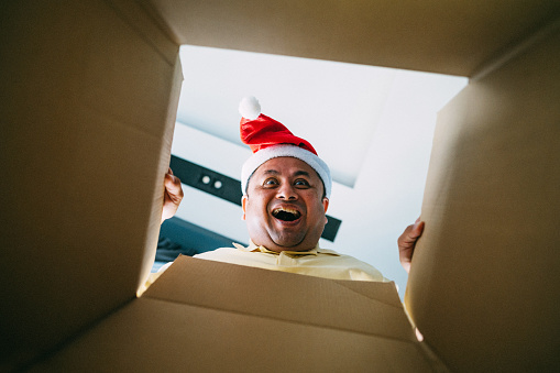 Excited guy smiling while opening present and looking inside. Portrait of guy opening carboard box while looking at camera. - Presents, delivery and holidays concept