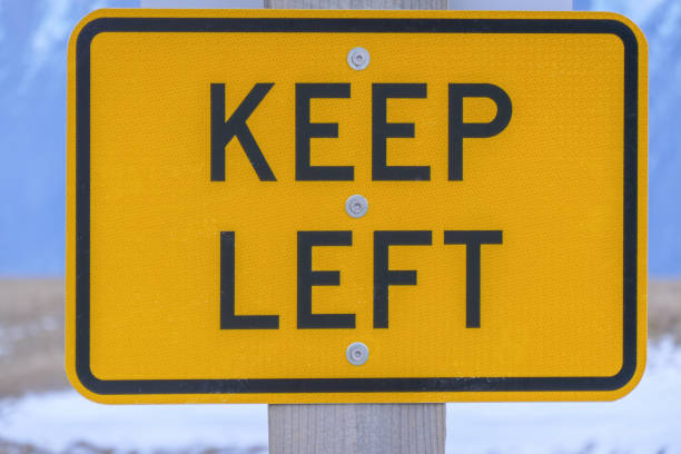 Keep Left Road sign reminding drivers to keep left on the road when leaving Round Hill ski field.  These reminders assist tourists who may be used to driving on the right side of the road.  In the background is snow and ice on the ground and the Southern Alps.  This image was taken on a cold, overcast day in early Spring. left wing politics stock pictures, royalty-free photos & images