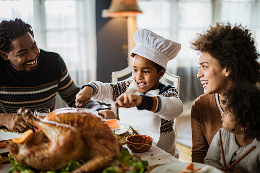 Happy African American small chef having fun while ready to eat whole Thanksgiving turkey during lunch with her family at dining table.