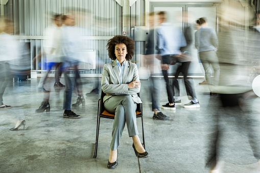 Young black businesswoman looking at camera while being surrounded by her colleagues who are walking in blurred motion.