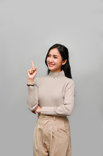 Portrait, Confident and beautiful millennial Asian female smiling, pointing up her finger, standing against the grey studio background. recommend, sharing ideas or opinion gesture.