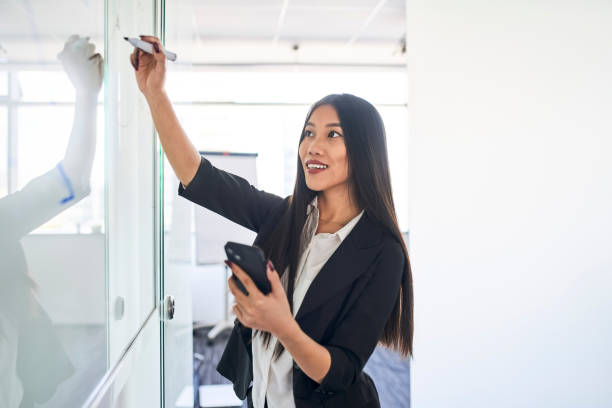 istock Asian businesswoman writing on whiteboard in office using mobile phone 1428954483