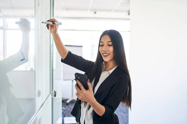 istock Young Asian businesswoman using her smartphone while writing on whiteboard in office 1428954608