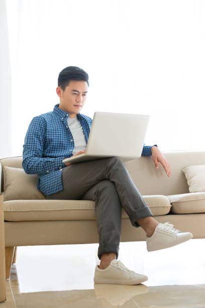 Casually dressed young man sitting on an indoor living room sofa using a laptop - stock photo Suitable for moving, gender emotions, interior decoration, interior design, housing rental, real estate, apartment rental and sale, home decoration, newlywed home ownership, urban white-collar rental, couple relationship, second-hand house renovation, weekend home relaxation, women's care, women's employment rights, print advertising remote work visas stock pictures, royalty-free photos & images