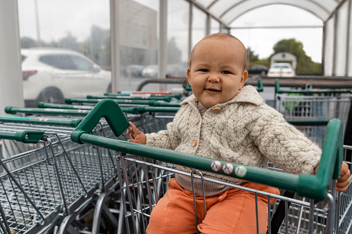 Medium shot of a baby boy who is sitting in a shopping trolley ready to go into a supermarket in the North East of England. He is smiling, looking at the camera.