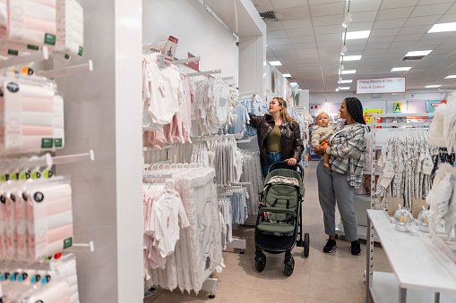 Wide shot of two woman shopping,  looking at baby clothing in the North East of England. One of the women is holding her baby while the other pushes a stroller.
