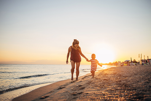 A mother and her child walking on the beach near the water in the sunset.