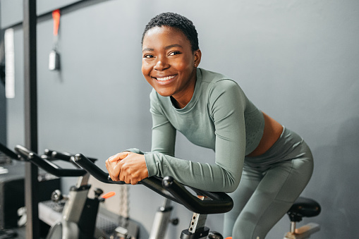 Black woman, exercise bike and gym fitness for energy workout, training and healthy wellness in cardio club. Portrait, smile and happy personal trainer on cycle machine for sports, power and strength
