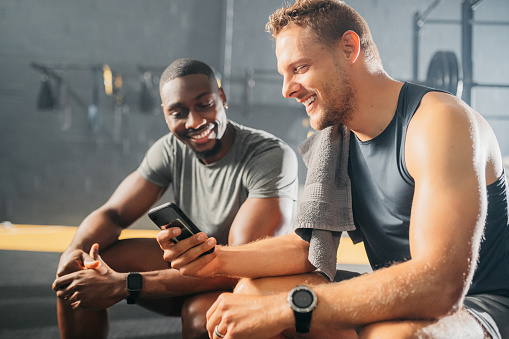 Men on their smartphone after a workout in the gym. Friends looking on the screen of a phone, smiling and laughing at a picture or video after exercising, training and doing fitness together