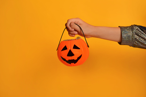 Orange pumpkin candy bucket or Halloween Jack o Lantern candy holder with sweets in female child hand on a yellow background with copy space.

Happy Halloween concept.