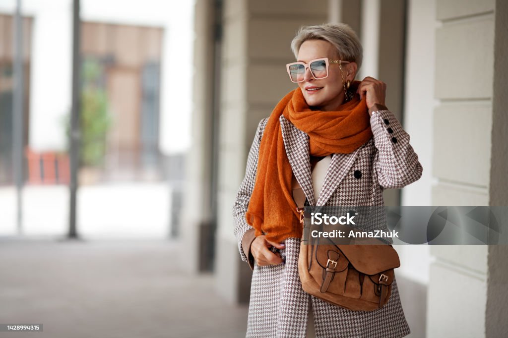 Smiling Woman Outdoor Portrait. Short blonde hair fashion model wears stylish clothes, double-breasted jacket, leather handbag, ochre knitted scarf and glasses. Fashion trend of autumn or spring Fashion Stock Photo