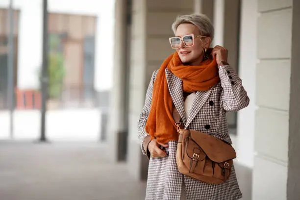 Photo of Smiling Woman Outdoor Portrait. Short blonde hair fashion model wears stylish clothes, double-breasted jacket, leather handbag, ochre knitted scarf and glasses. Fashion trend of autumn or spring