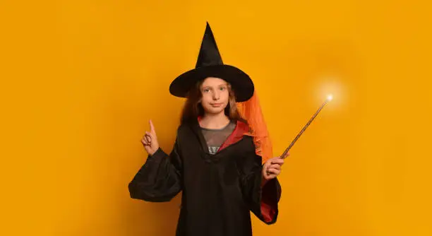 Cute Halloween child girl in wizard costume with luminous magic wand looking at camera and pointing index finger up on a yellow background.

Pre-teen girl holding finger up with glowing magic stick.
