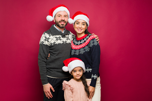 Happy smiling family celebrating the holidays wearing christmas sweaters and santa hats against a red background
