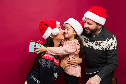 A young girl is wearing a santa hat and holding a large Christmas present, her parents and brother are sitting in the background also wearing santa hats