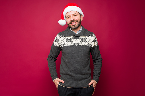 Hispanic handsome man wearing a christmas sweater and a santa hat enjoying the christmas holidays in front of a red background