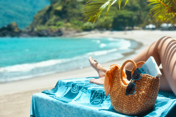 Woman legs lying on towel relaxing on summer holidays, near beach bag with a book and sunglasses on background of resort beach. stock photo
