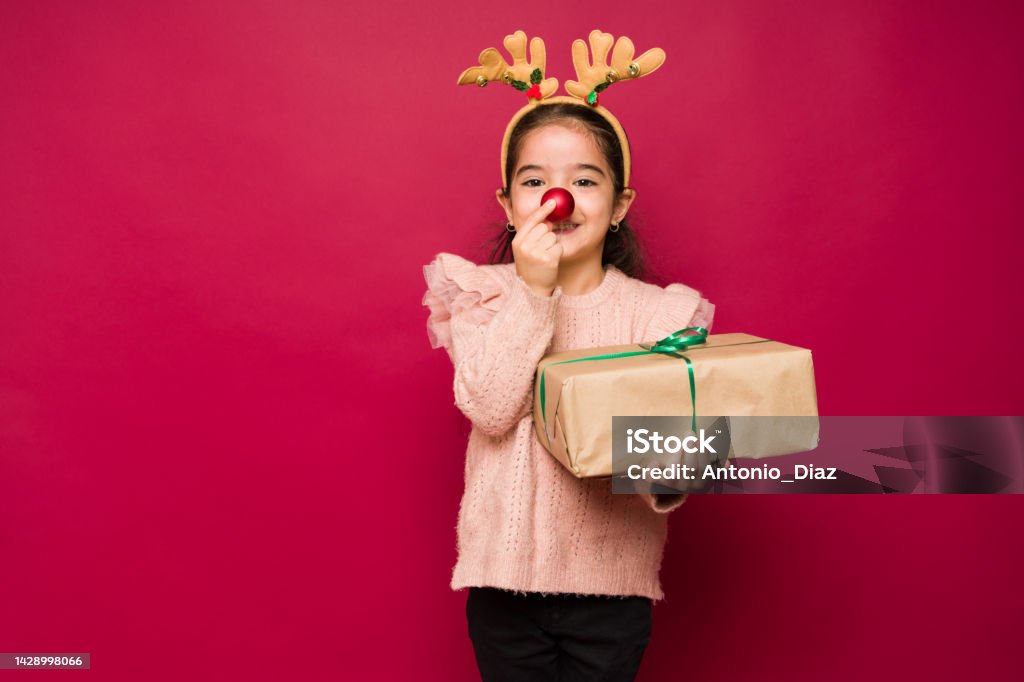 Adorable girl putting up a red reindeer nose for christmas Cute little kid playing with a reindeer nose and costume while enjoying the christmas holidays and presents Christmas Stock Photo