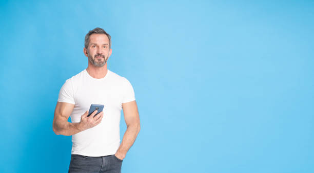 istock Handsome middle aged grey haired man 40s standing with with smartphone in hand wearing white t-shirt isolated on blue background. Mature fit man, healthy lifestyle concept 1429000627