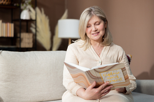 An elderly beautiful smiling woman holding a photo album sits on a comfortable sofa in the living room happily looking at family photographs of vacation memories.