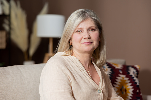 Portrait of beautiful smiling woman with blonde gray hair relaxing in living room on couch. Older mature complexion of elegant retired senior lady.