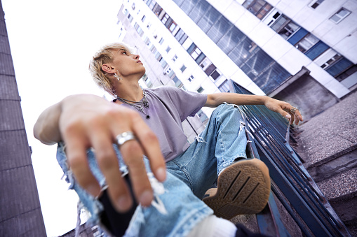 A teenage boy is sitting on a railing and chilling in an urban exterior surrounded by buildings.