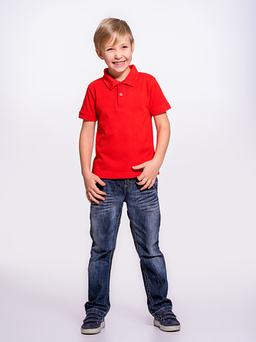 Young pretty boy posing at studio as a fashion model. Photo of a 8 years old kid. Full portrait of happy boy, isolated.  Portrait of white smiling kid in a red t-shirt and jeans. Boy in a full length