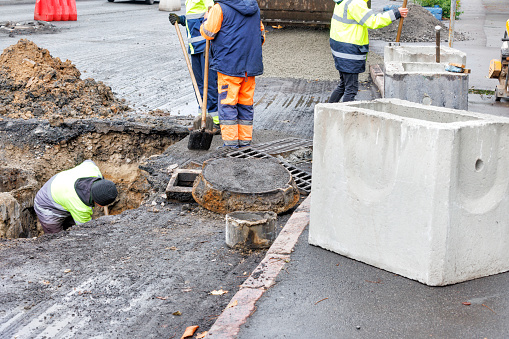 A group of road workers repair sewer storm hatches and change old concrete manholes for new ones on a roadway on an autumn day.