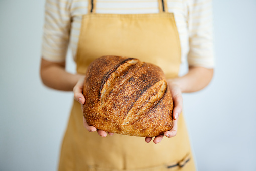 An anonymous woman in apron standing in a kitchen and holding freshly baked sourdough bread.