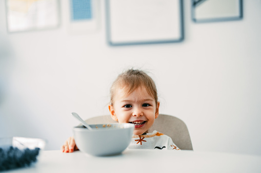 Portrait of smiling girl having fun while eating cereals with fruit for breakfast in the kitchen.