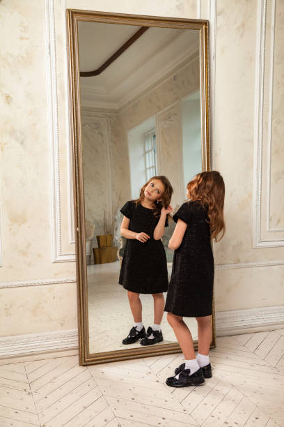 petite fille princesse de 6-7 ans aux cheveux bouclés, mannequin en élégante robe noire élégante au miroir dans le salon, regardant le reflet. jeune actrice à la mode à la maison. espace de copie - 6 7 years little girls child standing photos et images de collection