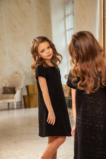 portrait petite princesse 6-7 ans blonde fille mannequin dans une robe élégante noire élégante au miroir dans le salon, regardant le reflet. jeune actrice à la mode à la maison. copier l’espace de texte - 6 7 years little girls child standing photos et images de collection