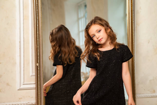 petite fille princesse de 6-7 ans aux cheveux bouclés, mannequin en élégante robe noire élégante au miroir dans le salon, regardant la caméra. jeune actrice à la mode à la maison. espace de copie - 6 7 years little girls child standing photos et images de collection