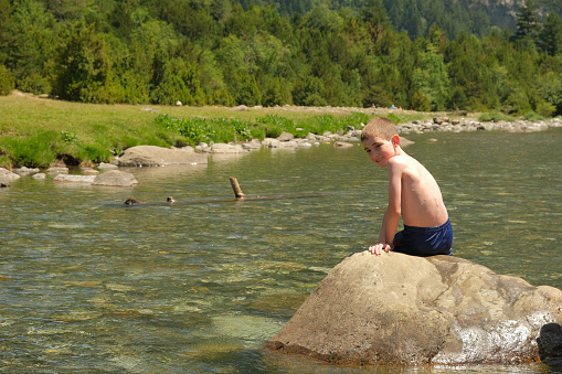 A boy is sitting on a rock in the river with his shirt off. He is surrounded by water, about to bathe. He is in the middle of a mountain, surrounded by green forest. It is a sunny and warm day.