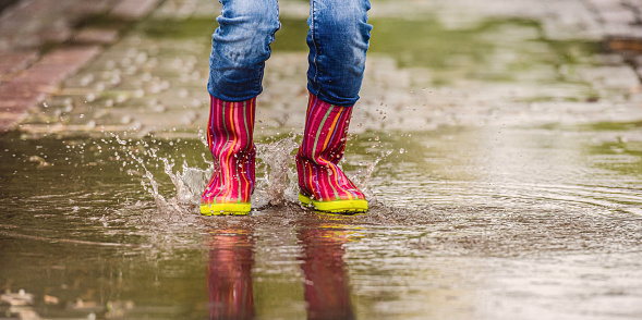 Girls legs in rubber boots making splashes from puddle on pavement, panoramic orientation