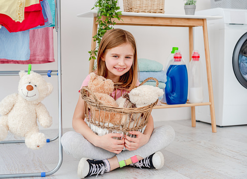 Smiling little girl holding plush toys in washing basket sitting on floor in bathroom