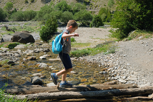 A boy is crossing a stream over some timbers. He is balancing to get across. He is in the mountains on a hot, sunny day. He is dressed in sports clothes with a backpack and hiking shoes. It is a general image and in profile.