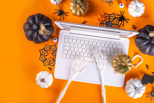 Funny Halloween workplace with laptop flat lay. Office corporation Halloween party invitation, blogger autumn holiday background. White laptop with Halloween party decor, orange background top view