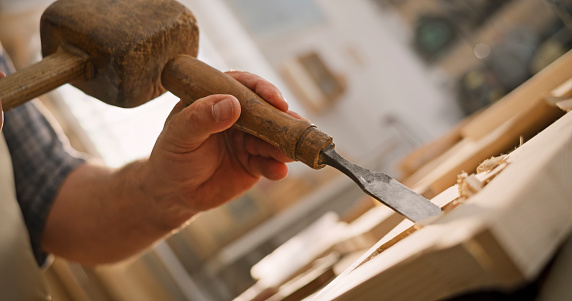 Close-up of male carpenter carving wood with chisel and mallet in workshop.